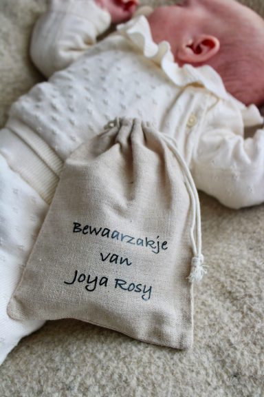 Een stoffen zakje met de tekst "Bewaarzakje van Joya" naast een baby in witte kleding.